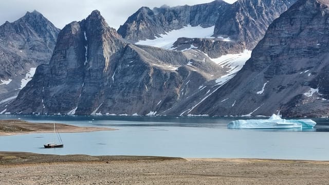 Peaceful landscape of Greenland fjord with iceberg and sailboat under rugged mountains.