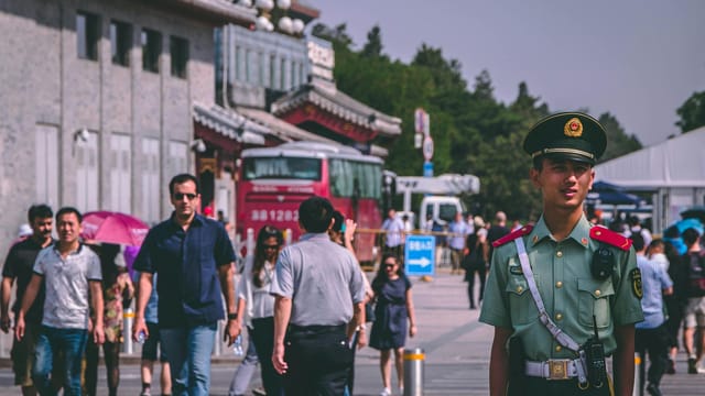 A Chinese police officer stands on a bustling street with people walking by on a sunny day.