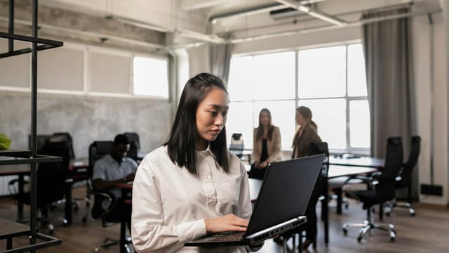 A focused woman using a laptop in a spacious, well-lit office environment with colleagues nearby.