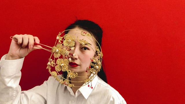 Elegant portrait of an Asian woman holding a golden ornament in front of her face with a vivid red background.
