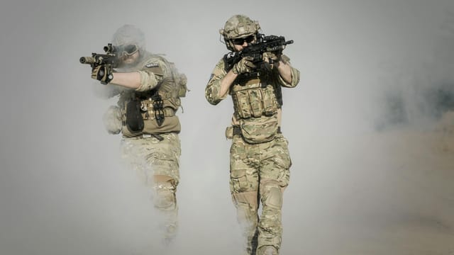 Two soldiers in camo uniforms with weapons advancing through a smoke-covered field, showcasing military action.