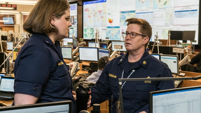 Adult and mature serious female colleagues in uniforms talking and looking at each other while standing near tables with computers behind stand with maps in modern office