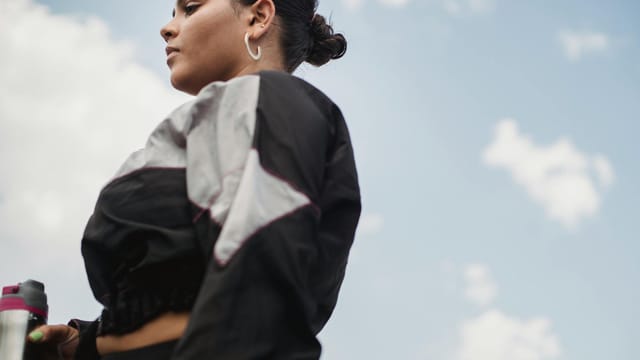 A woman poses confidently outdoors with a sports jacket against a blue sky and clouds.