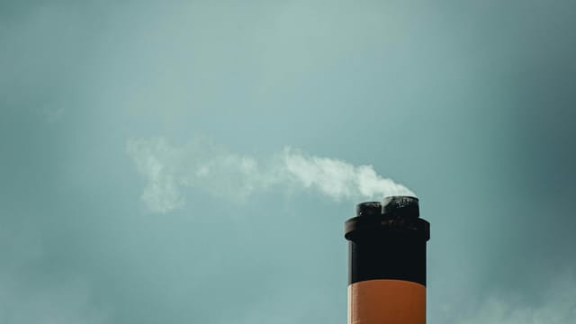 A tall chimney releasing smoke against a cloudy sky highlights industrial pollution.