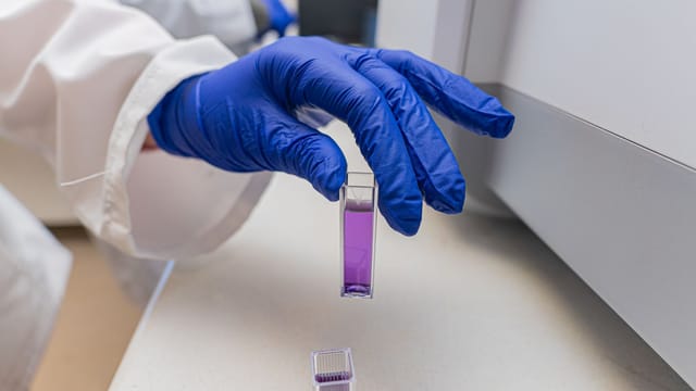 Close-up of a scientist's gloved hand handling a lab sample vial containing a purple liquid.