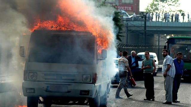 A van engulfed in flames on a city street with bystanders observing the scene.