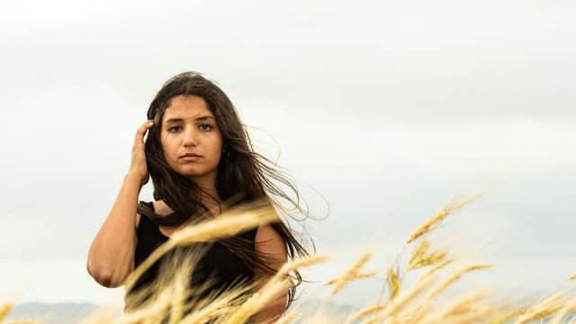 A contemplative woman standing in a windy field in Masyaf, Syria, captured in a moody atmosphere.
