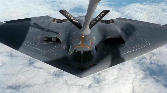 A stealth bomber aircraft receives aerial refueling mid-flight over clouds.