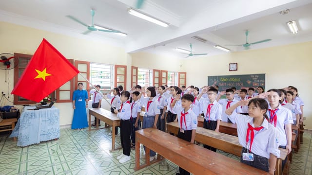 Students in uniform saluting the Vietnamese flag during a school ceremony in a classroom.