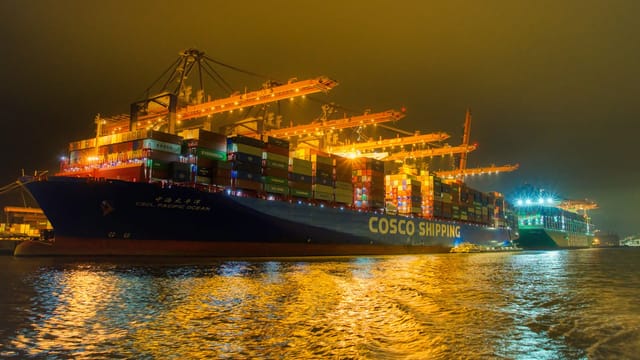 Illuminated container ship and cranes in Rotterdam harbor under overcast night sky.