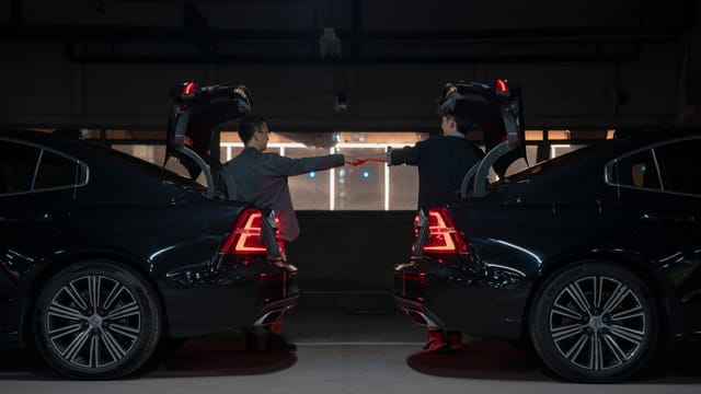 Two men in a dimly lit garage fist bumping between two cars with open trunks.