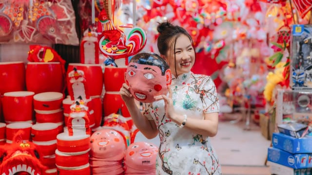 A smiling woman holds a colorful mask at a vibrant Asian market filled with traditional crafts.