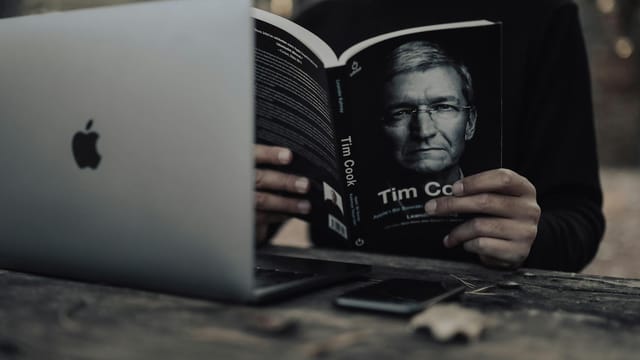 Unrecognizable ethnic male in warm clothes with opened laptop reading book and sitting outside at wooden table in autumn day