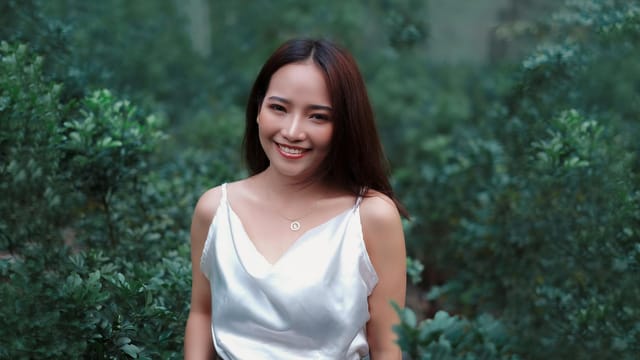 Smiling woman in a white top surrounded by lush green foliage outdoors.
