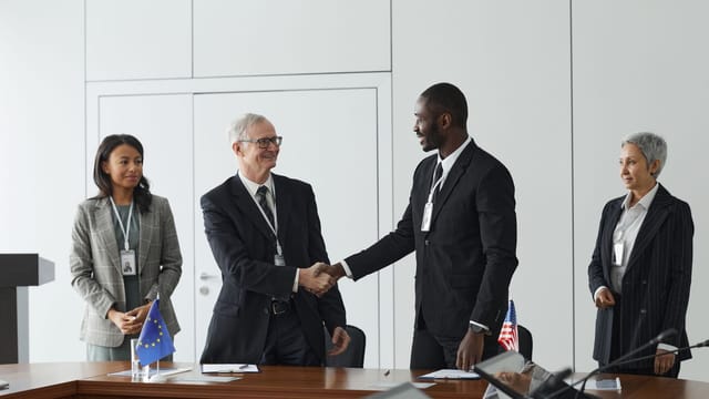 Business professionals shaking hands in conference room with flags signifying international agreement.
