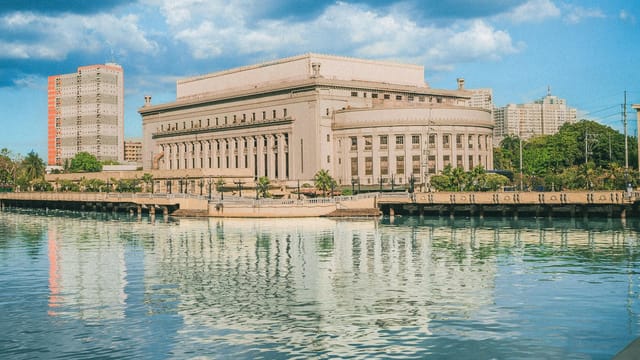The iconic Manila Post Office building reflecting on the Pasig River under a bright blue sky.