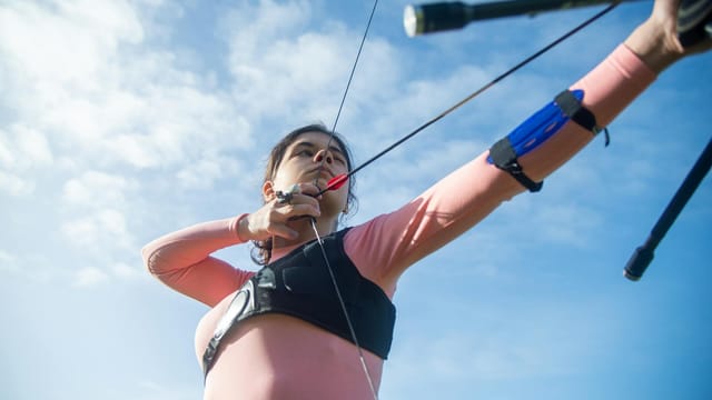 Woman focusing on archery with a recurve bow under a blue sky, highlighting precision and concentration.