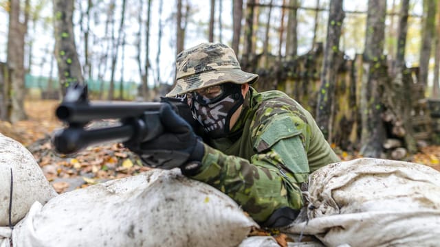 Man in camouflage uniform with mask aiming rifle in wooded area during training.