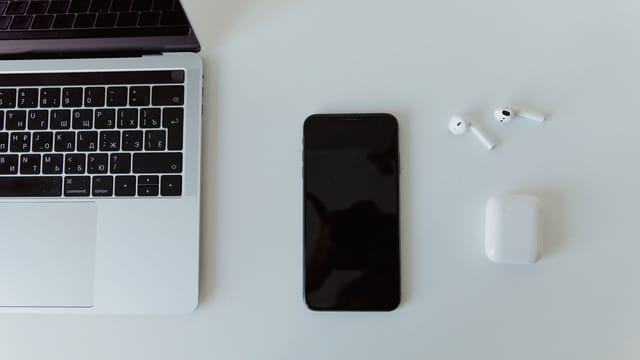 Top view of a laptop, smartphone, and wireless earphones on a white desk.