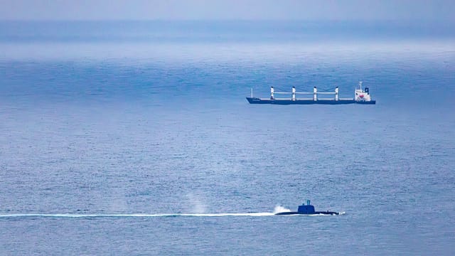 A cargo ship and submarine navigate the vast, open ocean under a clear sky.