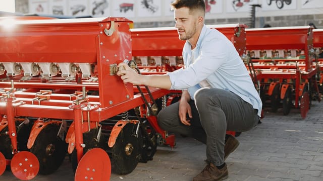 A young man examines red agricultural machinery outdoors on a sunny day.