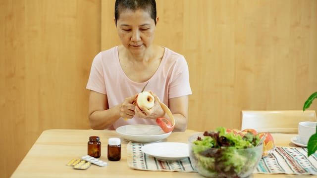 Middle aged ethnic female with short hair in t shirt peeling fresh apple and preparing salad while sitting at table with veggies and various medicine during remission