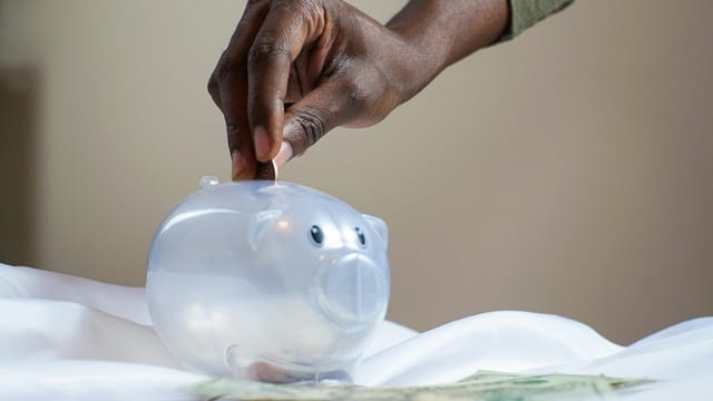 Close-up of a person's hand placing coins into a transparent piggy bank to save money.