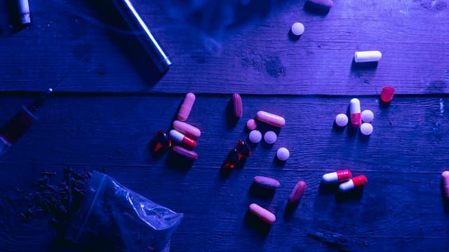 A flat lay of various drugs including pills, capsules, and a syringe on a wooden surface under moody blue lighting.