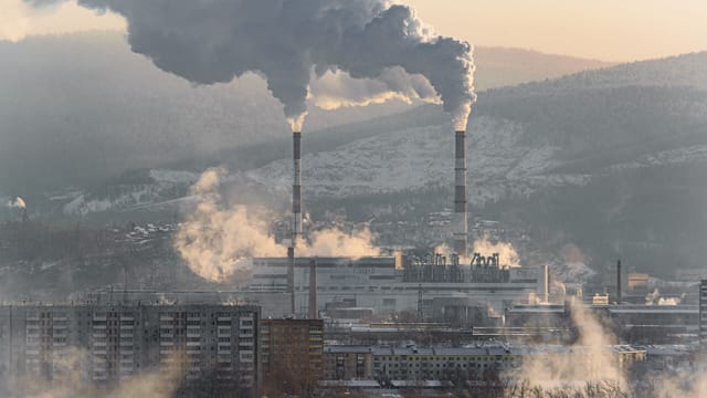 An industrial factory with chimneys emitting smoke, surrounded by snowy landscapes and buildings.