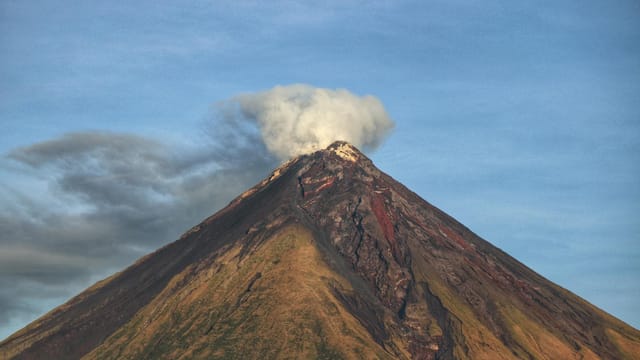 A dramatic image of an active Mayon Volcano emitting smoke under a clear blue sky.