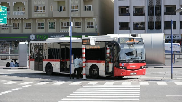 Public bus parked at a Dubai street bus stop with city buildings in the background.