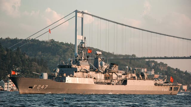 Military vessel with Turkish flags sailing under the Bosphorus Bridge in daylight.
