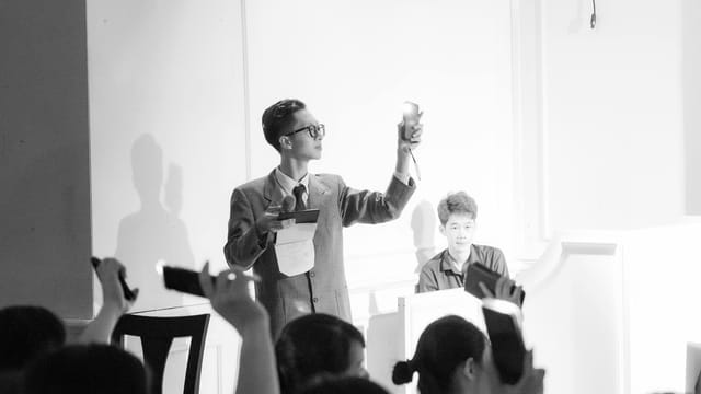Elegant black and white photo of an auction scene with a presenter and bidders raising phones.