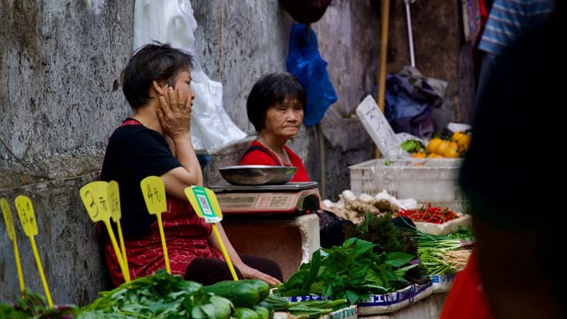Two women at an outdoor market stall with fresh produce and signs.