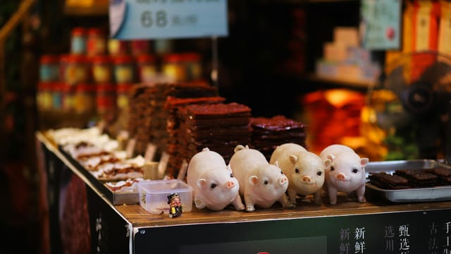 Market stall with assorted food items and ceramic pig figurines. Vibrant atmosphere.