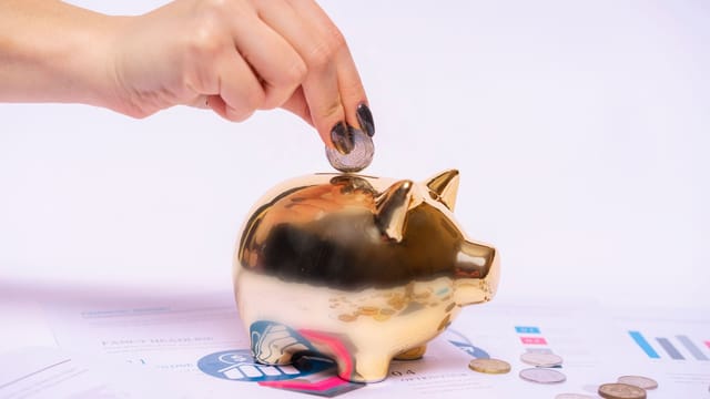 A hand places a coin into a golden piggy bank surrounded by financial documents, symbolizing savings.
