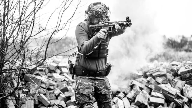 A soldier in full gear aims his rifle during a tactical training session on a rubble-strewn field.