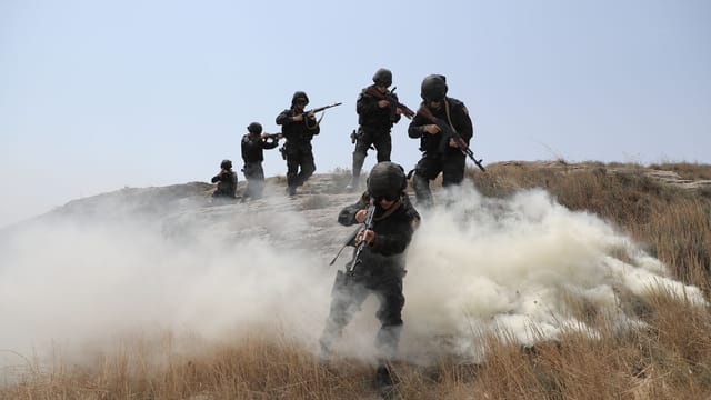 Soldiers advancing through a field with smoke during a training exercise.