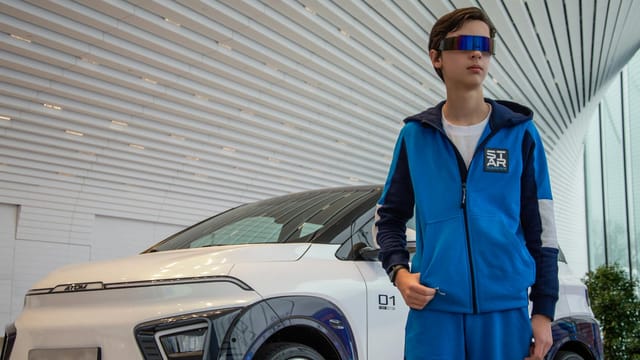 Teenager in blue tracksuit and futuristic glasses beside an electric car.