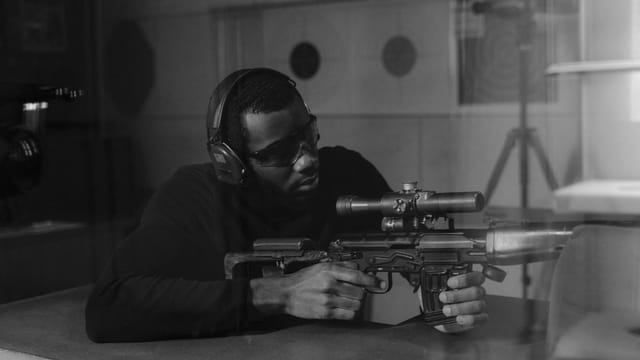 African American man with ear protection aiming a rifle indoors.
