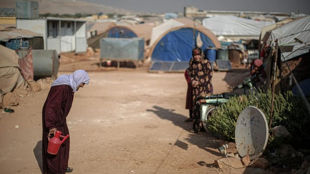 Syrian women engage in daily chores at a refugee camp under clear skies.