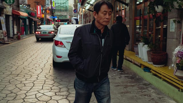 Asian man walking down a vibrant Suwon street lined with shops and cars.
