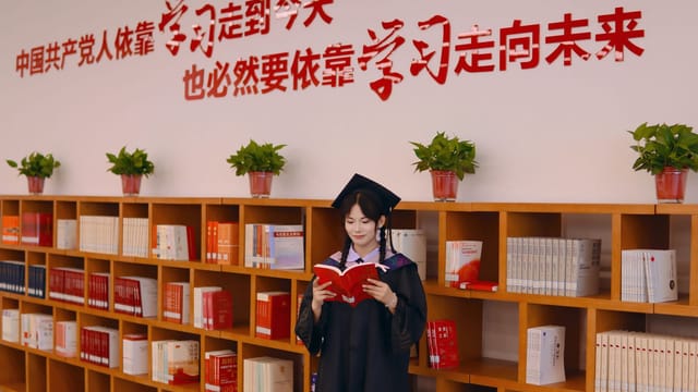 Female graduate in academic robe reading in a library with motivational Chinese text on the wall.