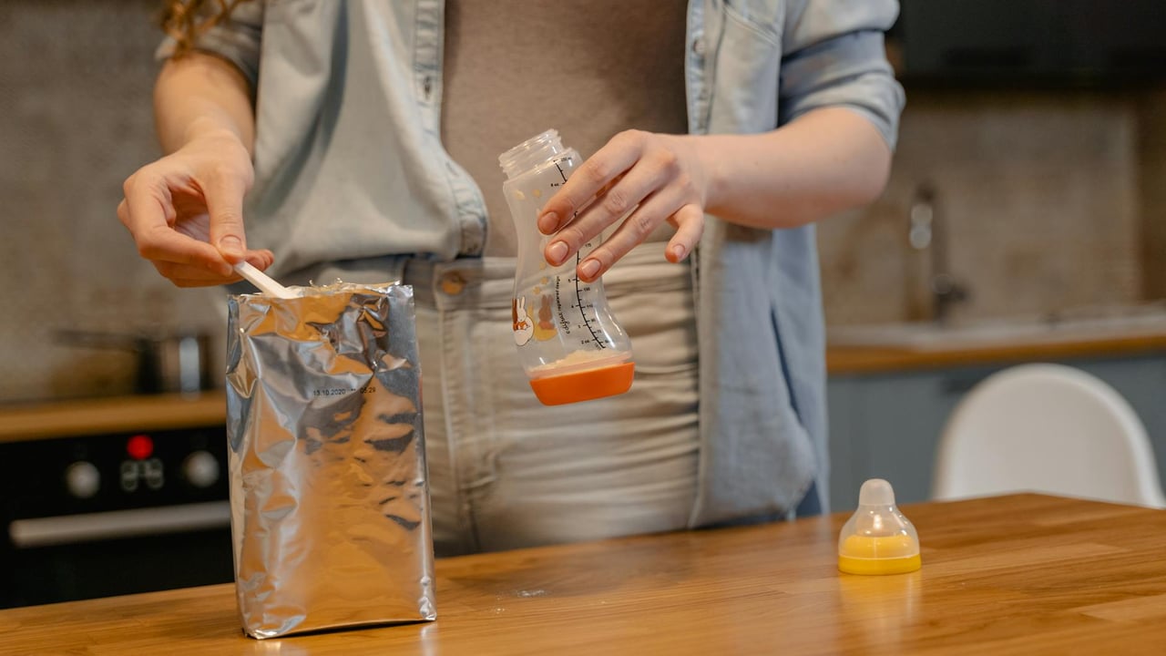 A parent prepares a baby bottle with formula on a wooden table in a cozy kitchen setting.