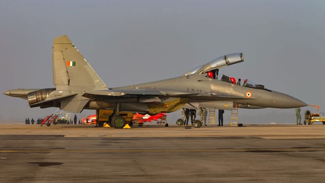 A military jet with open cockpit on a tarmac, surrounded by personnel and equipment.