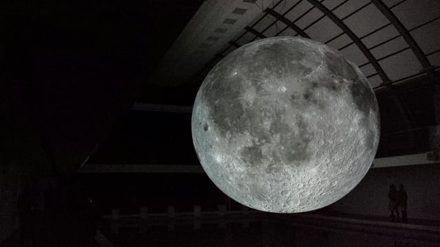 A captivating indoor display of a moon sculpture suspended in a museum, highlighting its spherical and cratered surface.