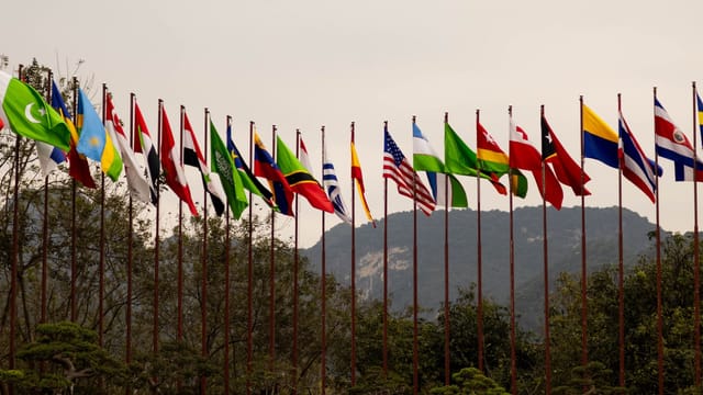 A display of various national flags on flagpoles set against a mountain backdrop, symbolizing global unity.