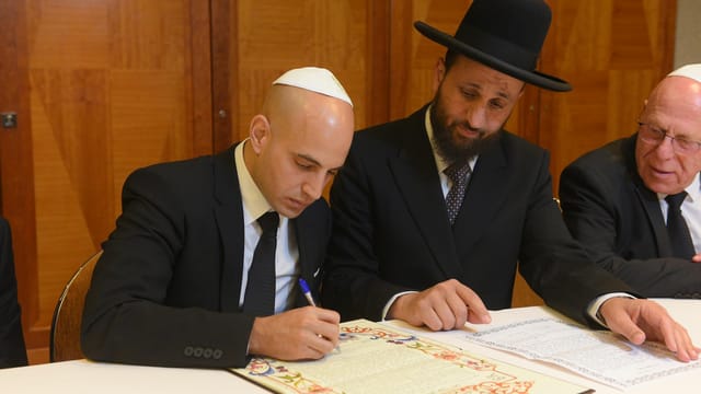Jewish men in suits sitting at a desk, reviewing and signing a document.