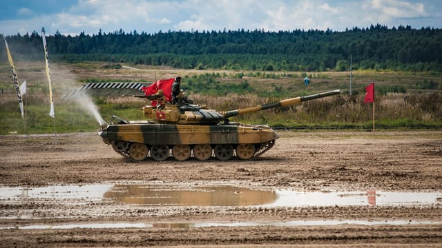 A military tank participating in a field training exercise with flags and muddy terrain.