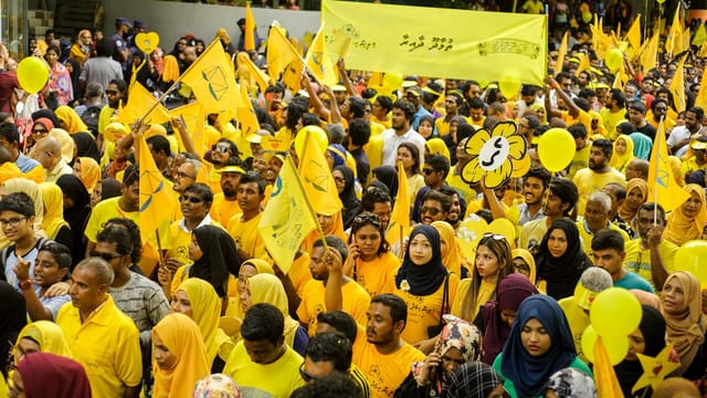 Crowd in yellow attends rally with flags, banners, and balloons, emphasizing unity.